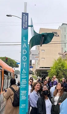 Light blue LA Dept of Transport bus stop pole with small mesh metal shade & light, with crowd looking up at it, some in disbe