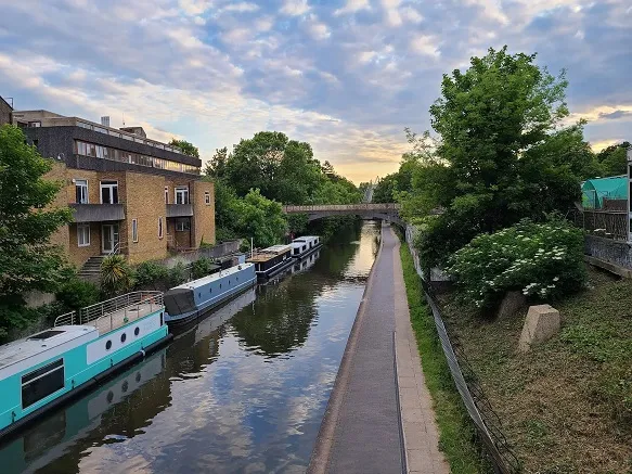 View of Regents Canal in London, with walking path on one side, and canal boats on the other, with houses behind them.